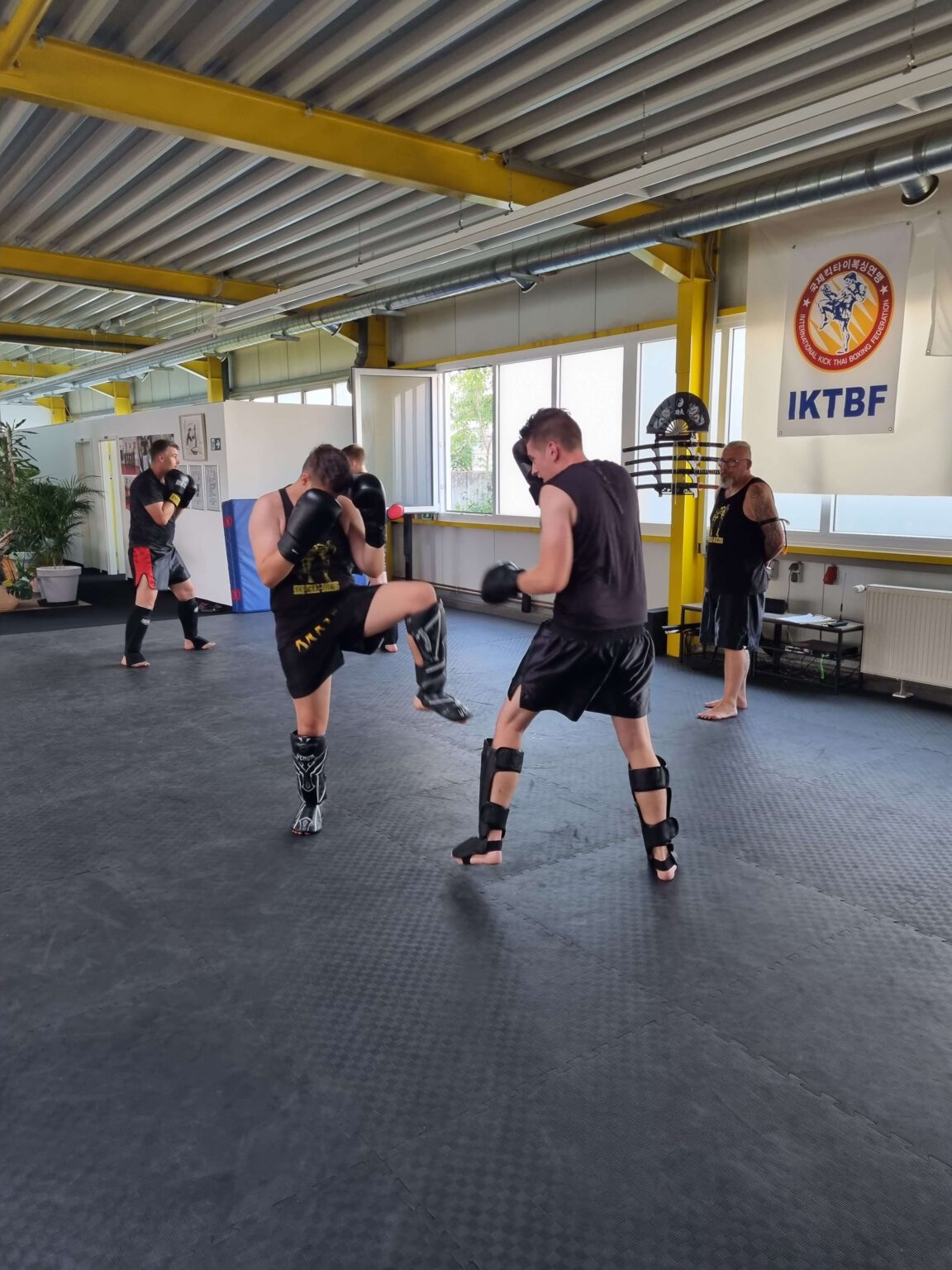 Mehrere Personen trainieren Kickthaiboxen in einem hellen Trainingsraum. Zwei von ihnen führen ein Sparring durch..