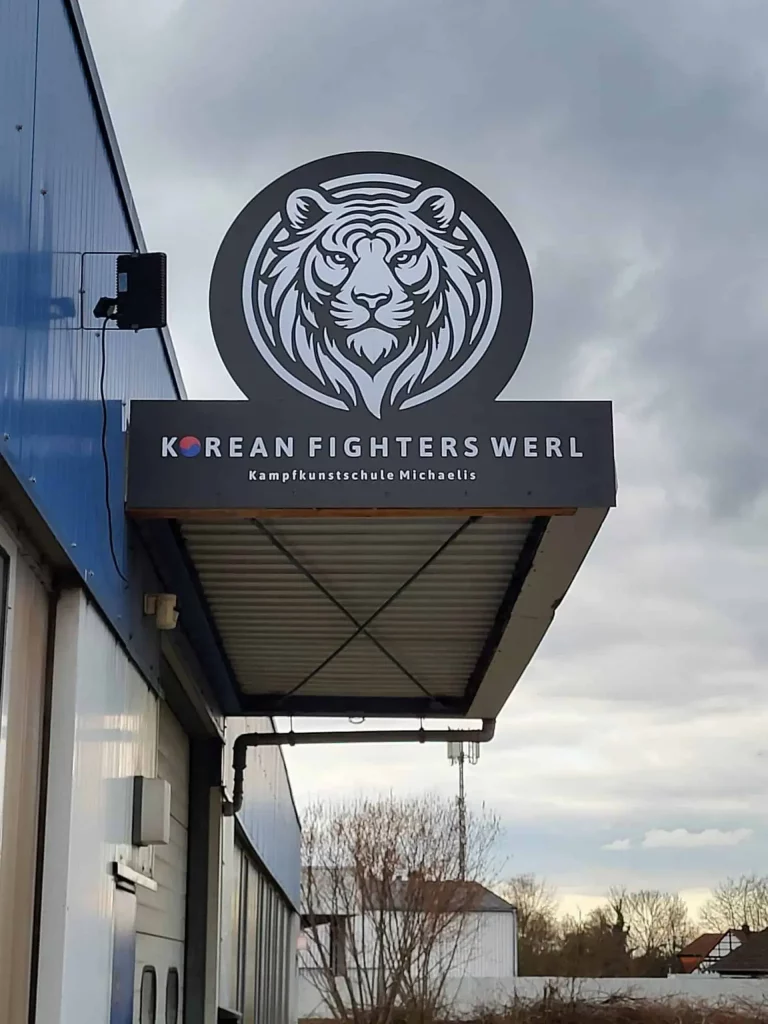Kampfkunstschule Korean Fighters Werl - Eingangsbereich - Aussenansicht Außenansicht der Kampfkunstschule Korean Fighters Werl: Ausleger-Schild mit Tigerkopf-Logo und Schriftzug „KOREAN FIGHTERS WERL – Kampfkunstschule Michaelis“ an einer blau verkleideten Hallenfassade, Himmel im Hintergrund.