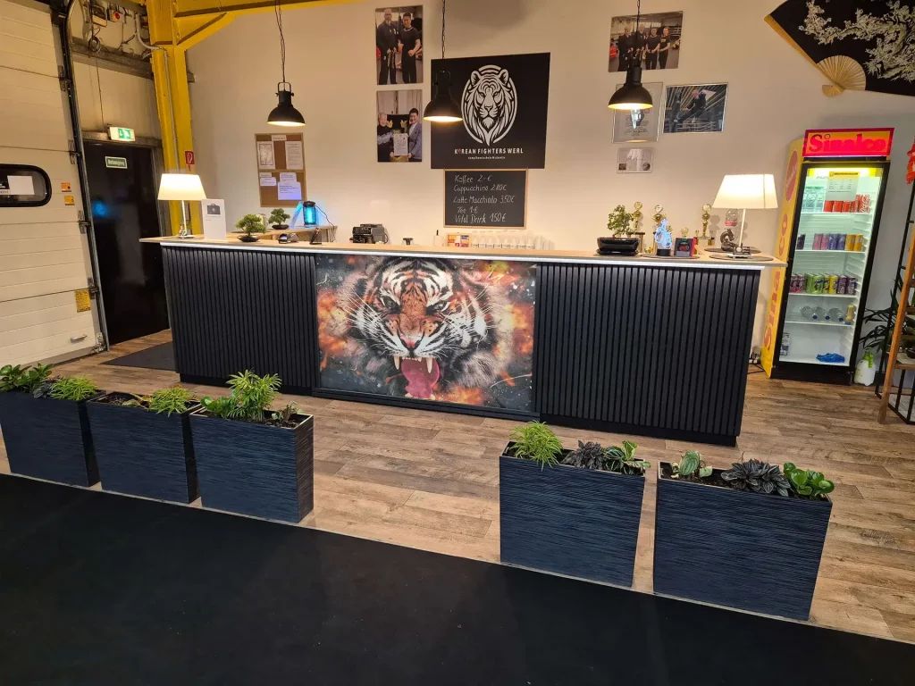 Empfangsbereich der Korean Fighters Werl mit breitem Tresen; vorne ein großes Tiger-Motiv, darüber Lampen und Wanddeko mit Fotos sowie Logo-Schild. Rechts ein Getränkekühlschrank, auf dem Tresen kleine Pokale und Deko – freundlicher Eingangs- und Servicebereich.