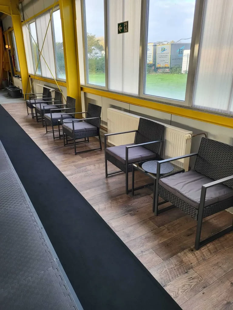 Kampfkunstschule Korean Fighters Werl - Sitzbereich Sitzbereich entlang der Fensterfront bei Korean Fighters Werl: mehrere dunkle Sessel mit Polstern und kleine Beistelltische auf Holzfußboden, Heizkörper unter den Fenstern und Fluchtweg-Schild – Wartezone direkt am Trainingsbereich.