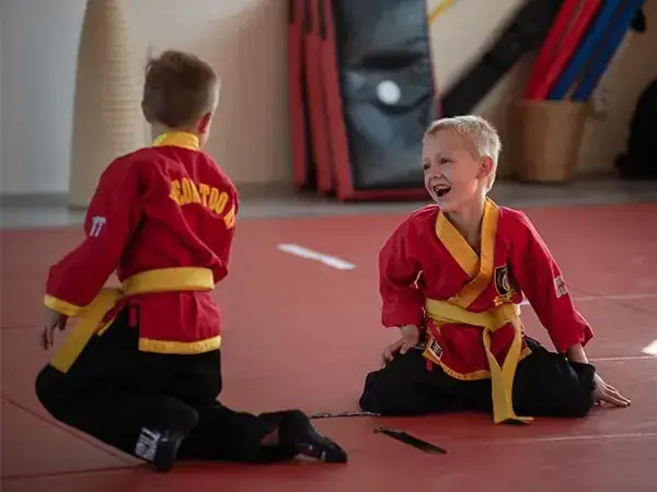 Kampfkunstschule Korean Fighters Werl - Kampfpsort in Werl - Kachel - Kyeok Too Ki Kids Korean Fighters Werl – Kampfsport in Werl: Zwei Kinder im roten Kyeok Too Ki-Anzug sitzen lachend auf der Matte und machen spielerische Koordinations- und Partnerübungen im Training.