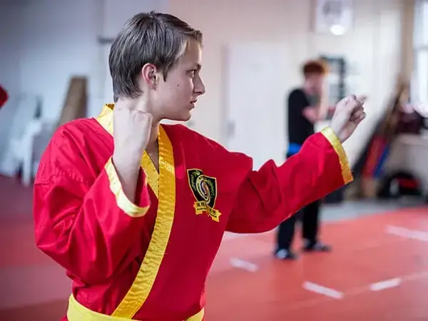 Kampfkunstschule Korean Fighters Werl - Kampfpsort in Werl - Kachel - Kyeok Too Ki und Kick-Thai Boxing für Jugendliche Korean Fighters Werl – Kampfsport in Werl: Jugendlicher im roten Kyeok Too Ki-Anzug steht in Kampfstellung mit erhobenen Fäusten und übt eine Schlagtechnik auf der Matte; weitere Trainierende unscharf im Hintergrund.